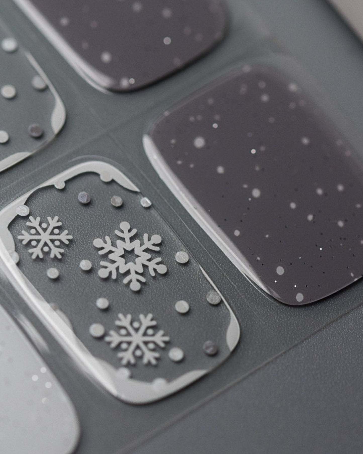 Nahaufnahme der Schneegrau UV Gel Wraps – kühle Grautöne, weiße Schneeflocken und dezente Golddetails für einen edlen Winterlook.

