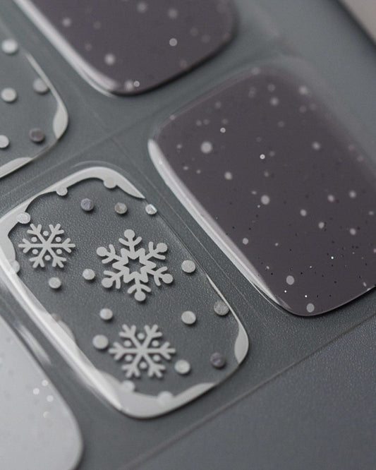 Nahaufnahme der Schneegrau UV Gel Wraps – kühle Grautöne, weiße Schneeflocken und dezente Golddetails für einen edlen Winterlook.
