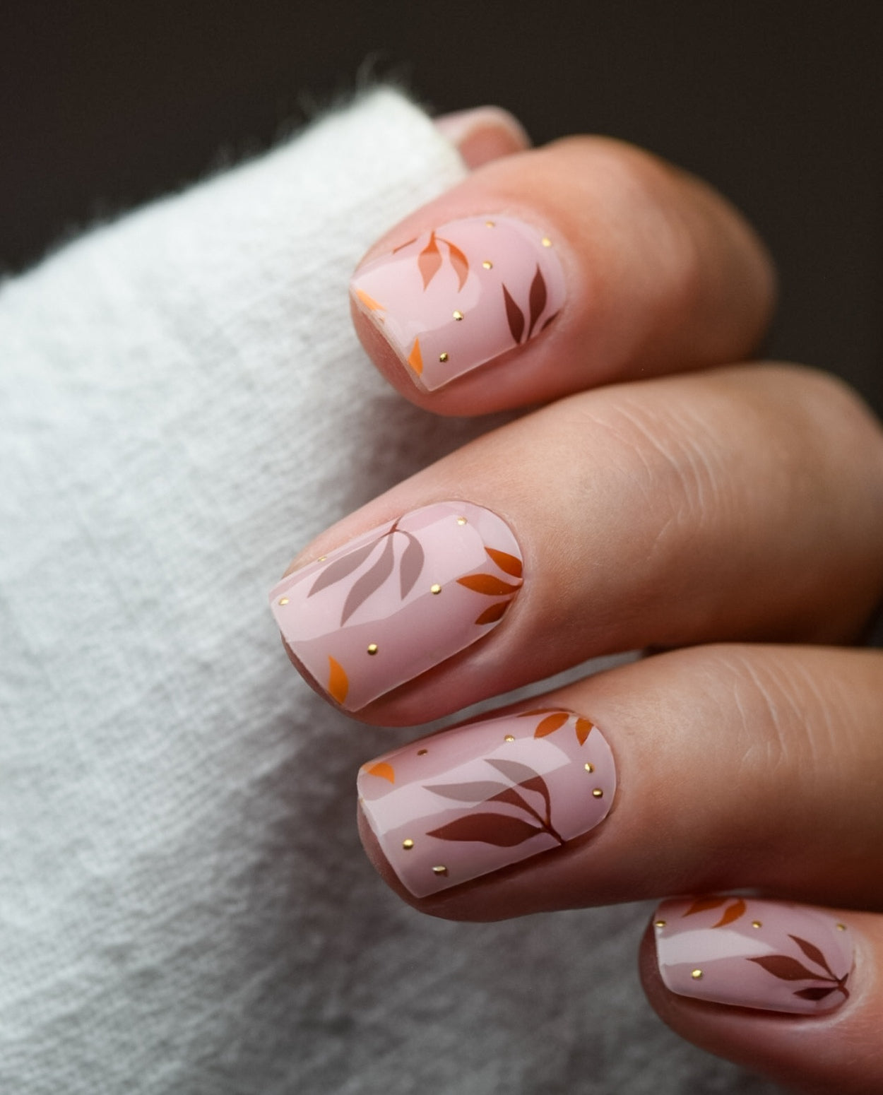 Hand mit UV Gel Wraps Cinnamon & Gold – zarter roséfarbener Hintergrund mit braun-goldenen Blättern, elegantes Herbstdesign.
