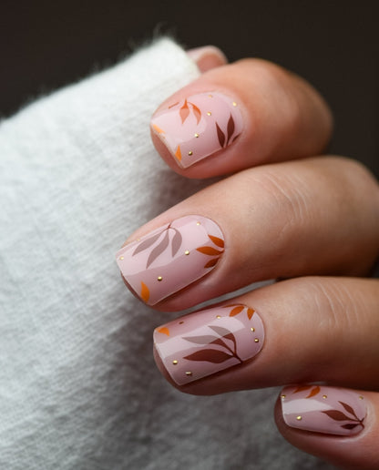 Hand mit UV Gel Wraps Cinnamon & Gold – zarter roséfarbener Hintergrund mit braun-goldenen Blättern, elegantes Herbstdesign.