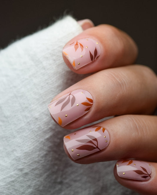 Hand mit UV Gel Wraps Cinnamon & Gold – zarter roséfarbener Hintergrund mit braun-goldenen Blättern, elegantes Herbstdesign.